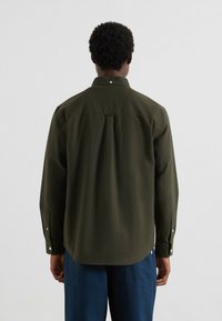 Uomo che indossa una camicia a maniche lunghe verde scuro con i bottoni e pantaloni blu, visto di spalle su uno sfondo chiaro e uniforme.