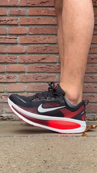 O persoană îmbrăcată cu pantofi de alergare Nike ZoomX negri și roșii, stând lângă un zid de cărămidă roșie pe un teren de beton.