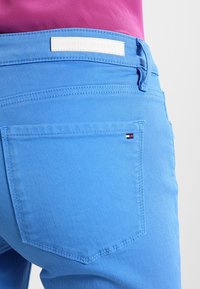 Pantalones de mezclilla azul con un bolsillo trasero que presenta un pequeño emblema tricolor. La cinturilla muestra una etiqueta blanca de Tommy Hilfiger.