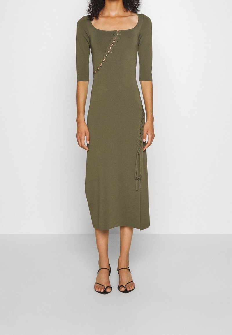 Femme portant une robe midi vert olive avec manches au coude et détails diagonaux à lacets, associée à des sandales noires à brides.