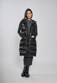 Schwarze Steppweste aus glänzendem Material mit Kapuze und Reißverschluss. Sie verfügt über Seitentaschen und ein knielanges Design, das über einem grauen Kleidungsstück getragen wird.