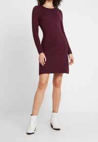 Abito in maglia bordeaux con maniche lunghe, scollatura rotonda e spacco laterale. Indossato con stivaletti bianchi alla caviglia con zipper sul retro.