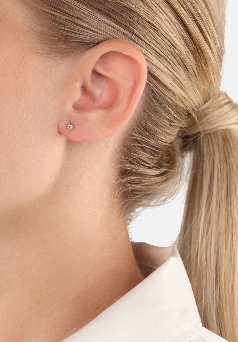 Primer plano de la oreja de una mujer con un pequeño pendiente de diamante redondo y el cabello rubio recogido en una coleta.