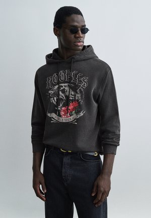 Young man wearing black sunglasses, dark hoodie with roaring tiger and red roses graphic, and dark jeans against plain light background.
