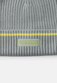 Szary prążkowany beanie z żółtym paskiem na mankiecie oraz niebieską metką z białym napisem "Calvin Klein".