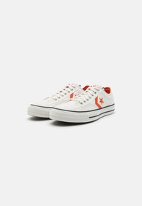 Zapatillas de lona blancas con acentos rojos, que presentan un logo de estrella, cordones blancos y una suela de goma texturizada con rayas negras.