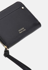Schwarze Leder-Geldbörse mit goldenen Beschlägen, Reißverschluss und eingeprägtem "GUESS Made in Italy"-Logo. Verfügt über einen Handgelenkriemen und Kartenfächer.
