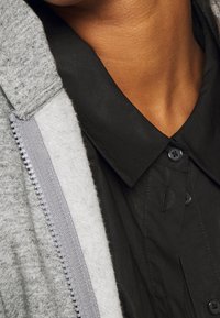 Gros plan d'une personne portant une chemise noire à col sous une veste polaire grise zippée.