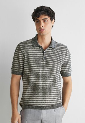 Ung mand med krøllet hår iført en grå og hvidstribet kortærmet polo-shirt og lysegrå bukser, står op ad en ensfarvet hvid væg.