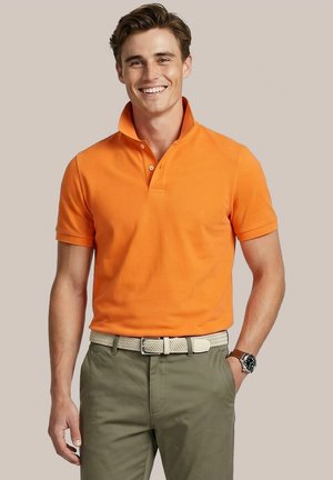 Lachende man met bruin haar, gekleed in een oranje poloshirt, een beige geweven riem, olijfgroene broek en een horloge met leren band.