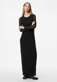 Černé maxi šaty s dlouhými průhlednými rukávy, kulatým výstřihem a splývavým střihem, spárované s robustními černými kotníkovými botami.