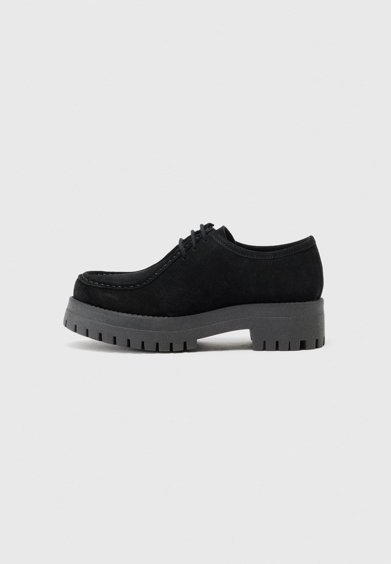 Chaussure à lacets en daim noir avec une semelle épaisse en caoutchouc gris à rainures. Présente un bout arrondi et un détail de couture minimal. Vue de côté.