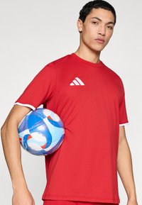 Κόκκινη αθλητική μπλούζα με λευκές λεπτομέρειες, με απλό σχέδιο και στρογγυλή λαιμόκοψη. Μια μπλε μπάλα ποδοσφαίρου με κυκλικά μοτίβα κρατιέται κάτω από το μπράτσο.