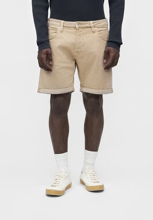 Mężczyzna w beżowych podwiniętych krótkich spodenkach, czarnej koszulce z długim rękawem, białych skarpetkach za kostkę i białych trampkach stoi na jednolitym tle.