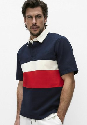 Mies, jolla on tummat hiukset ja parta, pukeutunut tummansinivalko-punaiseen raidalliseen lyhythihaiseen poolopaitaan, seisoo yhdellä kädellä taskussa.