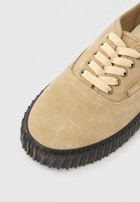 Baskets en daim beige avec des lacets marron clair, dotées d'une semelle en caoutchouc noire texturée et de détails de couture subtils. Aucune marque visible.