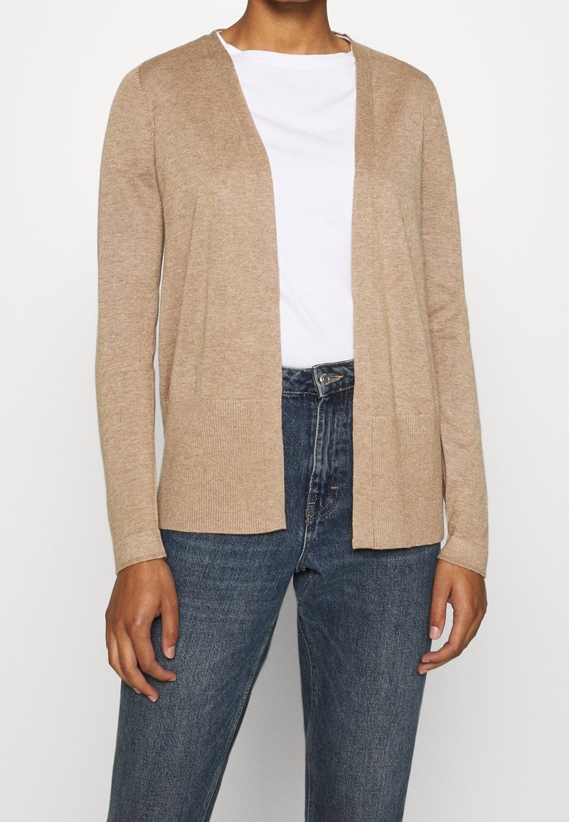 Beige cardigan with long sleeves, open front, and two front pockets. Made from a soft material, paired with a white t-shirt and blue jeans.