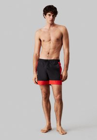 Costumi da bagno neri e rossi con vita elastica, cordino e orlo rosso a contrasto. Tessuto leggero, design di lunghezza media.