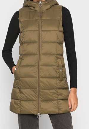 Osoba ubrana w długą, oliwkowozieloną pikowaną bezrękawnik z suwakiem z przodu i kieszeniami po bokach, na czarnym, prążkowanym topie z długim rękawem.