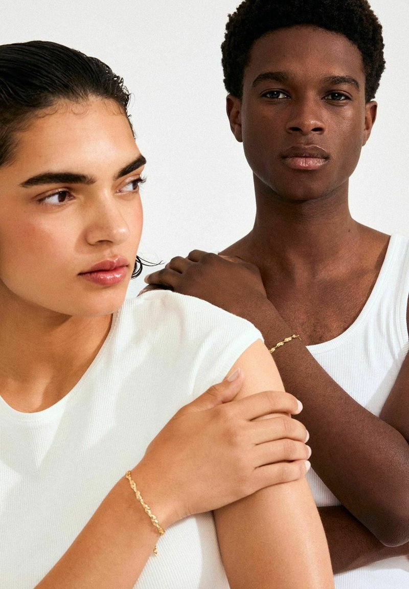 Kaks modellid esitavad kuldseid käevõrusid. Naine kannab valget keha järgiistuvat tippu ja mees valget tanktoppi, taust on neutraalse tooniga.