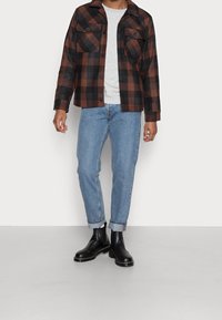 Czarna i brązowa kraciasta flanelowa kurtka z dwoma kieszeniami na piersi, zestawiona z jasnoniebieskimi dżinsami i czarnymi botkami na szarym tle.