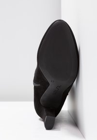 Bottine en daim noir à bout pointu avec un talon bloc, semelle tournée vers l'avant contre un mur et un sol blancs formant un angle.