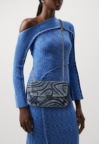 Abito blu a righe con maniche lunghe e design off-shoulder. Borsa trapuntata blu e grigia con tracolla a catena, caratterizzata da un motivo astratto e chiusura nera.