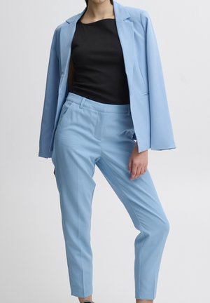 Femme portant un pantalon de costume bleu clair et un blazer assorti sur un haut noir, debout avec une jambe légèrement pliée.