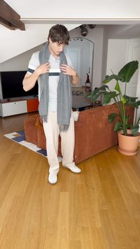 Sciarpa grigia drappeggiata attorno al collo, misto cotone, con bordi frangiati. Maglietta grigia chiara, pantaloni larghi color crema e sneakers bianche. Ambientazione interna.