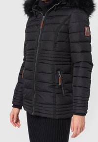 Chaqueta negra acolchada con capucha de piel sintética, costuras horizontales, cierre frontal con cremallera y bolsillos laterales, con detalles en cuero marrón.