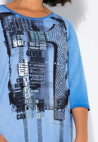 T-shirt en coton bleu à manches courtes, avec un motif de texte graphique en noir et argent, doté d'une texture en points et d'accents brillants.