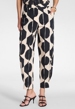 Donna che indossa pantaloni beige con audaci motivi geometrici neri e sandali neri con tacco a cinturino, mani nelle tasche.