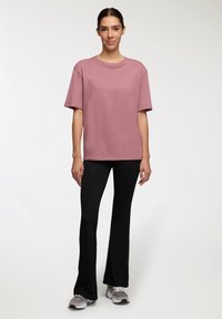 Jasnoróżowa koszulka z krótkim rękawem o luźnym kroju, w połączeniu z czarnymi spodniami dzwonami i szarymi butami sportowymi, stojąca na neutralnym tle.