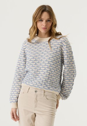 Frau mit cremefarbenem Pullover mit blauem Wellenmuster und beiger Hose steht vor hellem, einfarbigem Hintergrund und schaut nach vorne.