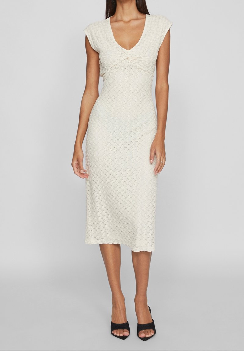Femme portant une robe midi texturée crème avec des manches courtes et un décolleté en V torsadé, associée à des talons ouverts noirs.