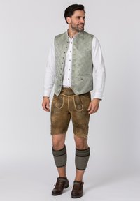 Grüner gemusterter Weste über einem weißen Hemd, kombiniert mit braunen Lederhosen mit dekorativer Stickerei und grauen Beinstulpen. Braune Schuhe.