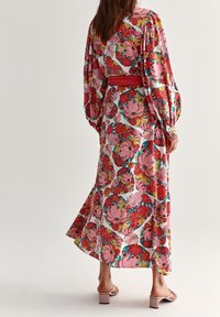 Vestido maxi floral con mangas largas, flores rosas y rojas sobre un fondo blanco, cintura ceñida con un cinturón rojo, dobladillo fluido.