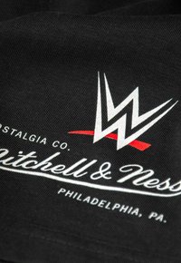 Schwarzer Stoff mit einem weißen WWE-Logo und roten Akzenten, daneben der Text "Mitchell & Ness" in Weiß und "Philadelphia, PA" darunter.