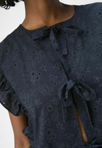 Blouse floral sombre avec des manches volantes, présentant une ouverture en forme de trou de clé et des liens décoratifs. Le matériau a un imprimé texturé subtil avec des découpes.
