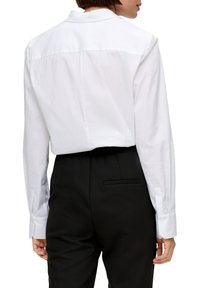 Camicia bianca a maniche lunghe con colletto a punta, dotata di uno yoke posteriore e polsini abbottonati, abbinata a pantaloni neri.