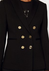 Blazer doppiopetto nero realizzato in tessuto testurizzato; presenta bottoni dorati con emblemi dettagliati e sottili accenti neri sulle tasche.