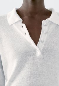 Weißes Strick-Poloshirt mit strukturierter Stoffqualität, ausgestattet mit einem klassischen Kragen, einer Dreiknopfleiste und kleinen Knopfdetails auf der linken Seite.