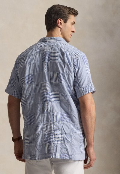 Άνδρας που φοράει μπλε και λευκό πουκάμισο με κοντά μανίκια και σχέδιο πατς, γυρισμένος με την πλάτη, με λευκό παντελόνι και καφέ ρολόι χειρός στο αριστερό του χέρι.