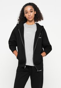 Felpa nera con zip e grande cappuccio, dotata di due tasche frontali, abbinata a una maglietta grigia con il marchio "PEGADOR" e pantaloni della tuta neri.