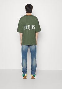 T-shirt vert surdimensionné avec le mot « PEQUS » en blanc dans le dos, associé à un jean bleu clair et des baskets multicolores.