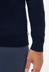 Marineblauer Strickpullover mit gerippten Bündchen und Saum. Etikett auf der linken Seite sichtbar. Die glatte Textur deutet auf ein weiches Material hin.