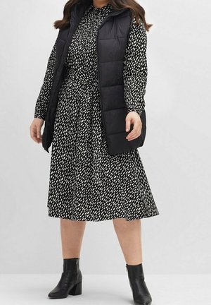 Gilet imbottito nero sopra un vestito con motivo bianco e nero e vita aderente. Stivaletti neri completano il look.