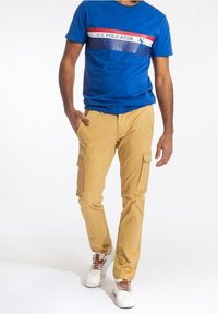 Blå kortärmad t-shirt med logotyp och randiga detaljer, kombinerad med beige cargobyxor med stora sidofickor och vita sneakers.