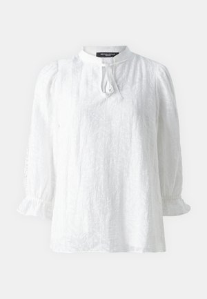 Blouse blanche à manches longues avec un col montant, tissu texturé et poignets à volants. Présente une fermeture à nouer sur le devant.