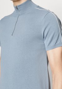 Kortärmad, ljusblå polo-skjorta med hög krage och dragkedja i kvartslängd. Har subtilt texturerad tyg och vit accentstickning.